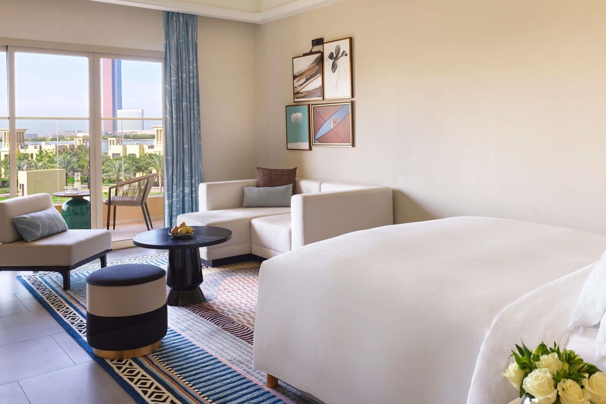 A hotel room with a large bed, two sofas, a small table, and a view of the cityscape.