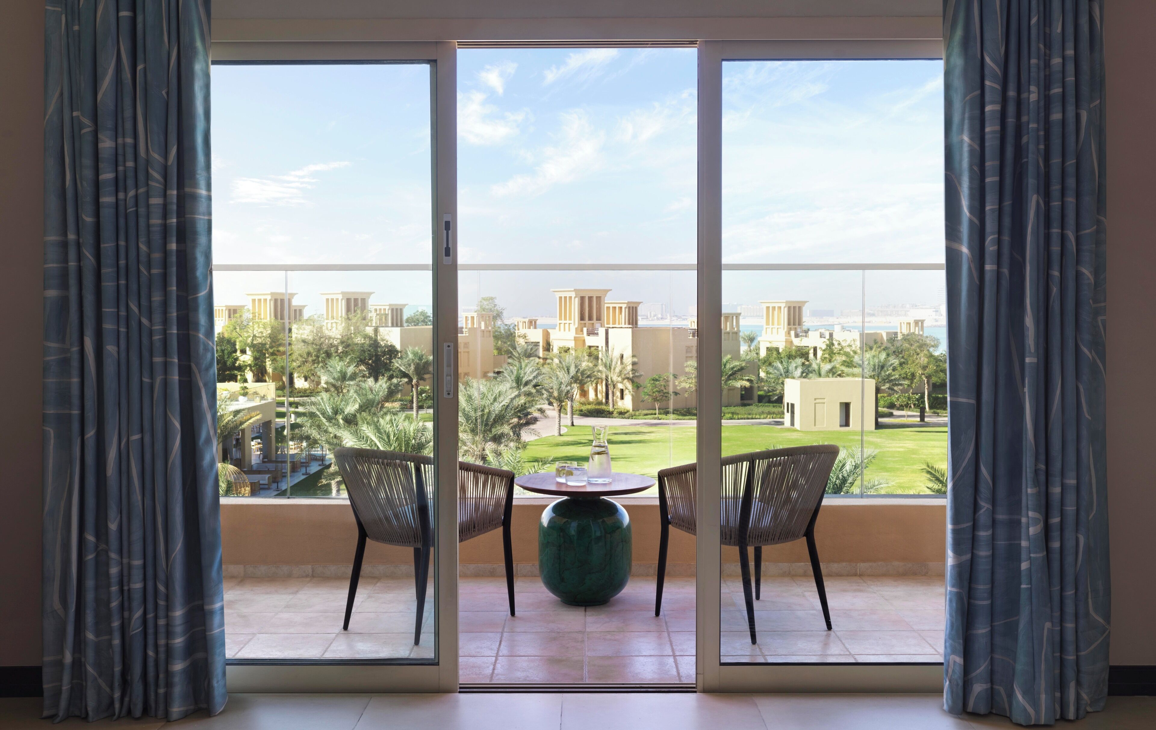 A balcony with two wicker chairs, a green vase, and a view of a residential area with palm trees and buildings.