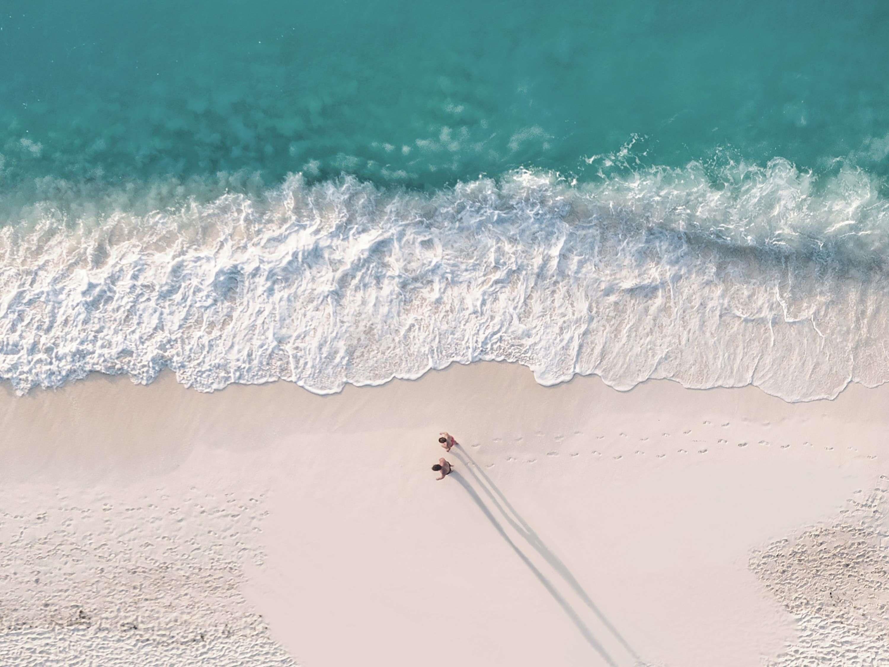 An aerial view of a beach with waves approaching the shore, a clear sky, and a single person walking along the beach.
