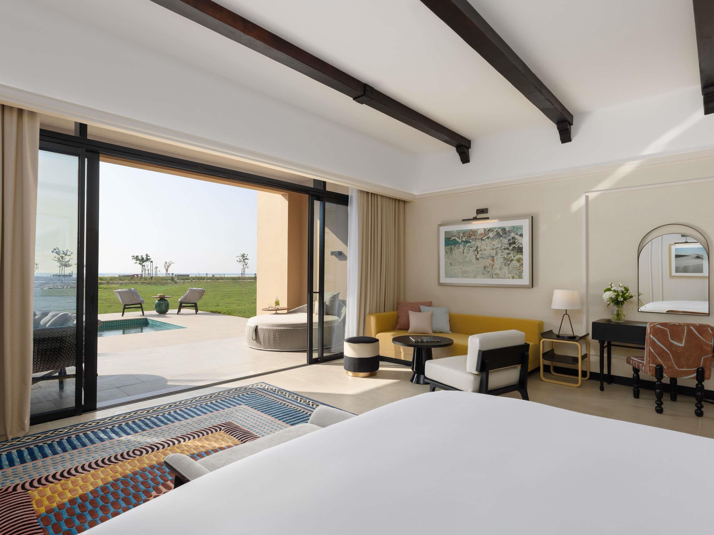 A hotel room with a large bed, a sofa, a small table, a chair, a desk, a lamp, a mirror, and a view of the beach.