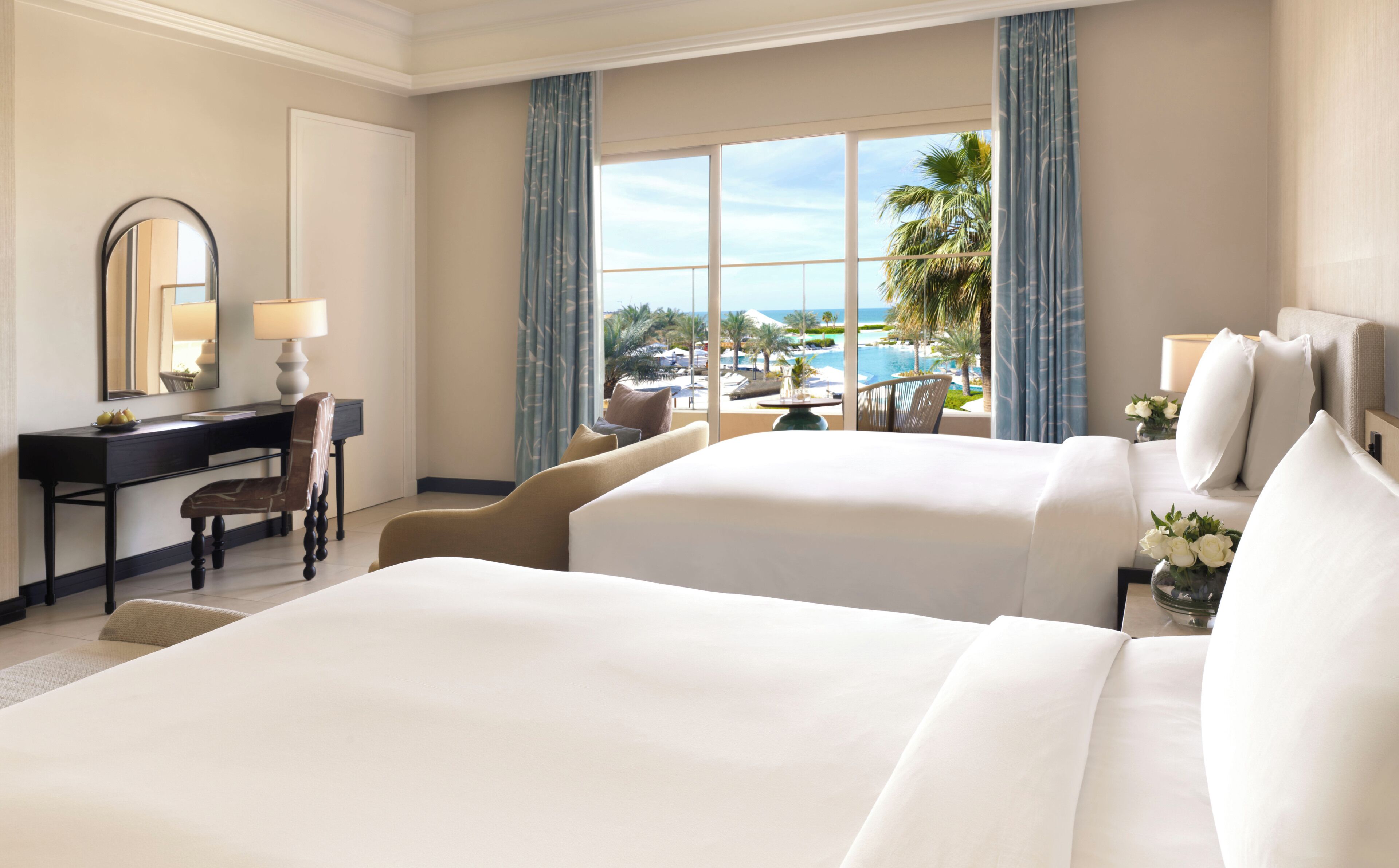 A hotel room with two beds, a desk, a chair, a mirror, and a view of a pool and palm trees.