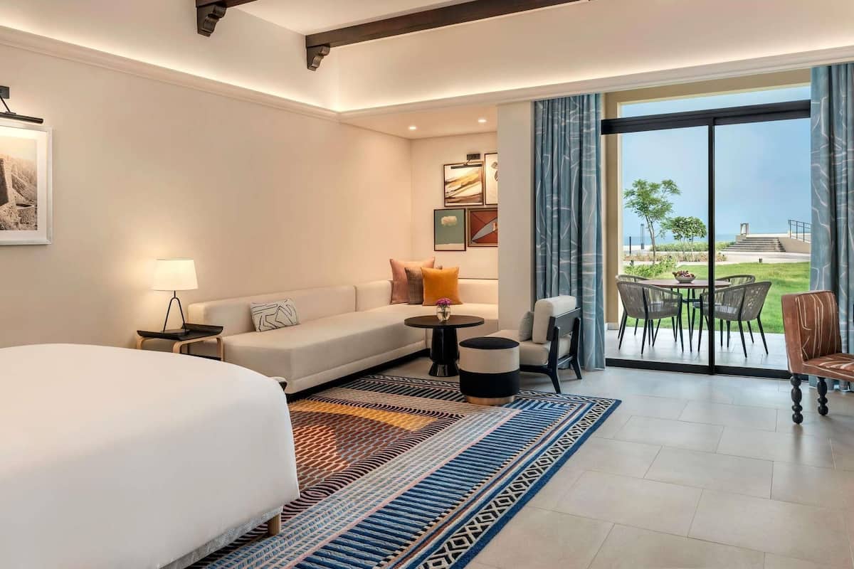 Un cuarto de hotel con una cama grande, un sofá, una mesa de comedor con sillas, una mesita de noche con lámpara y vista al exterior.