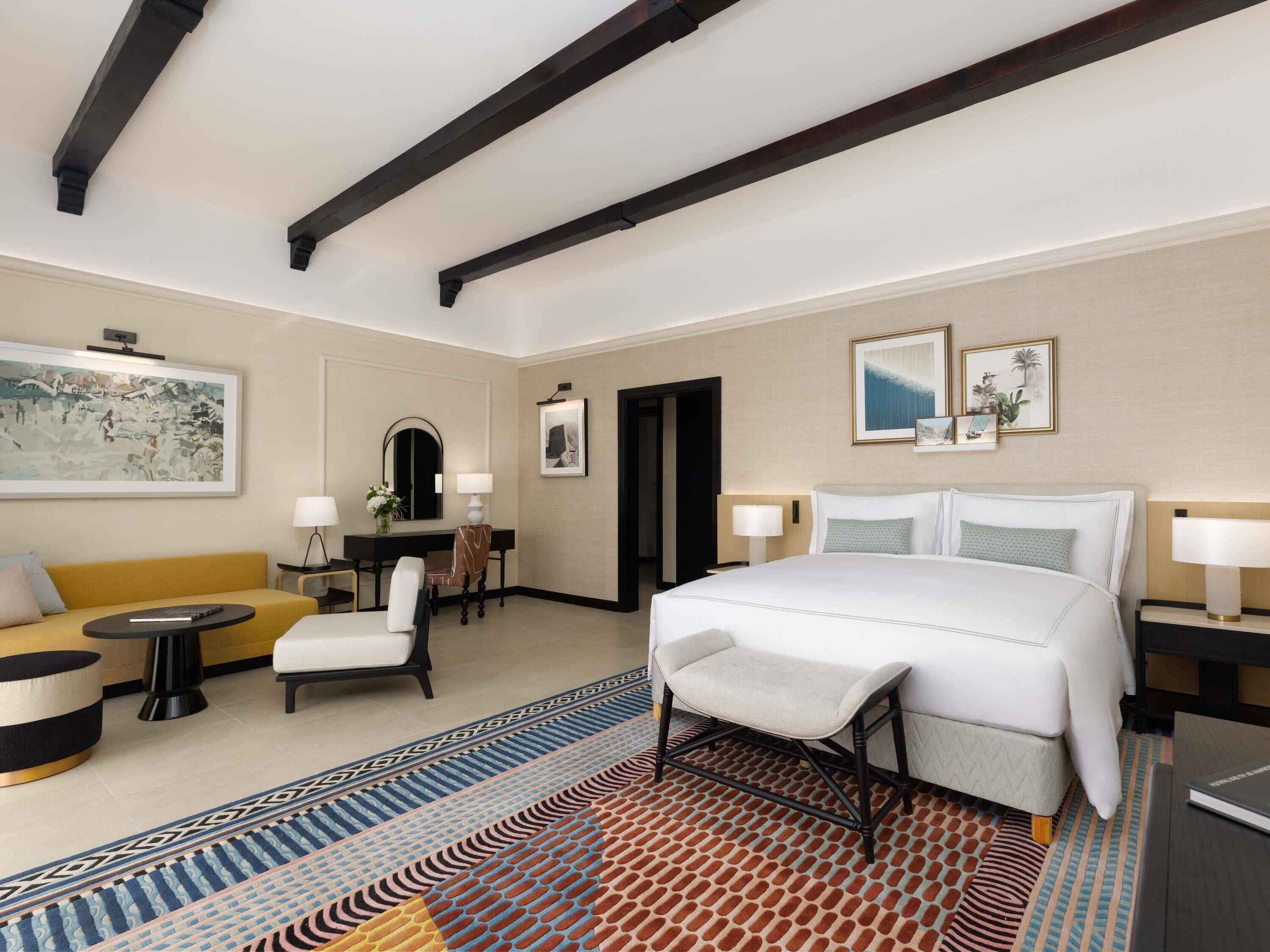 Une chambre d’hôtel avec un grand lit, un canapé, un bureau et une chaise. On y trouve deux lampes, un miroir et des tableaux encadrés accrochés au mur.