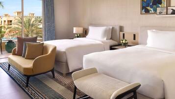 A hotel room with two beds, a sofa, a chair, a small table with a vase of flowers, and a view of palm trees and buildings.