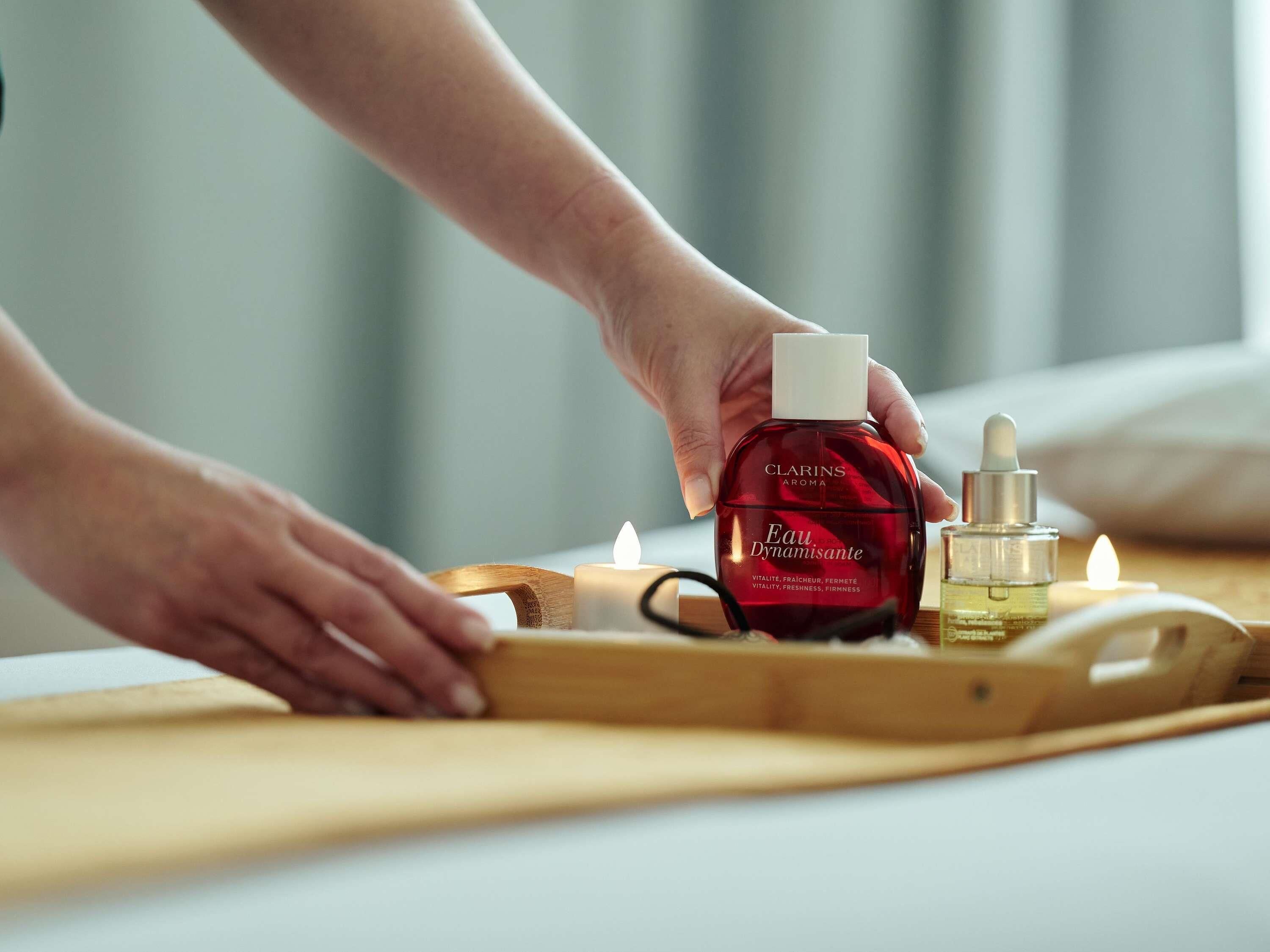 A hand holding a bottle of Clarins Eau Dynamisante on a tray with candles and a dropper bottle.
