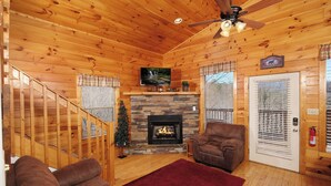L’intérieur chaleureux d’un chalet avec une cheminée en pierre, des murs en bois, un ventilateur de plafond et un téléviseur fixé au-dessus de la cheminée.