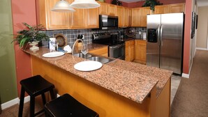 Una cocina con encimeras de granito, electrodomésticos de acero inoxidable, gabinetes de madera y un taburete de bar.