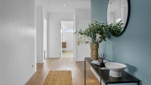 A hallway with a wooden floor, a woven rug, a vase with white flowers, a black-framed mirror, and a white basin on a black console table. - Penthouse in the City (Christchurch)
