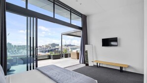 A modern hotel room with a large bed, a bench, a flat-screen TV, and a city view through large windows. - Penthouse in the City (Christchurch)