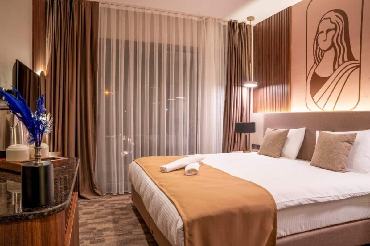 Ein Hotelzimmer mit einem großen Bett, einem Nachttisch, einem Spiegel und Blick auf die Stadt durch das Fenster.