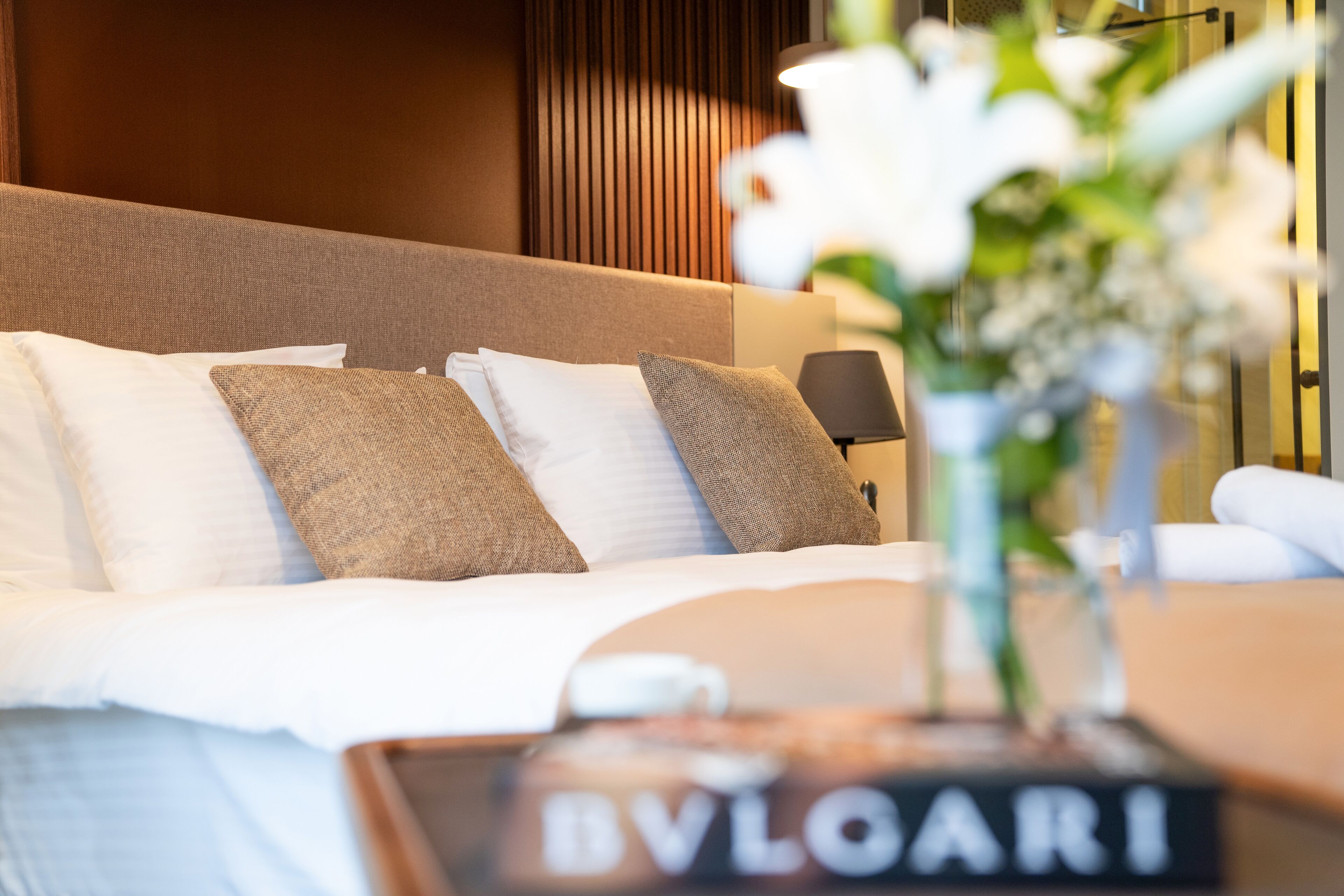 Una cama bien hecha con ropa de cama blanca y beige, una mesita de noche con lámpara y un jarrón con flores.