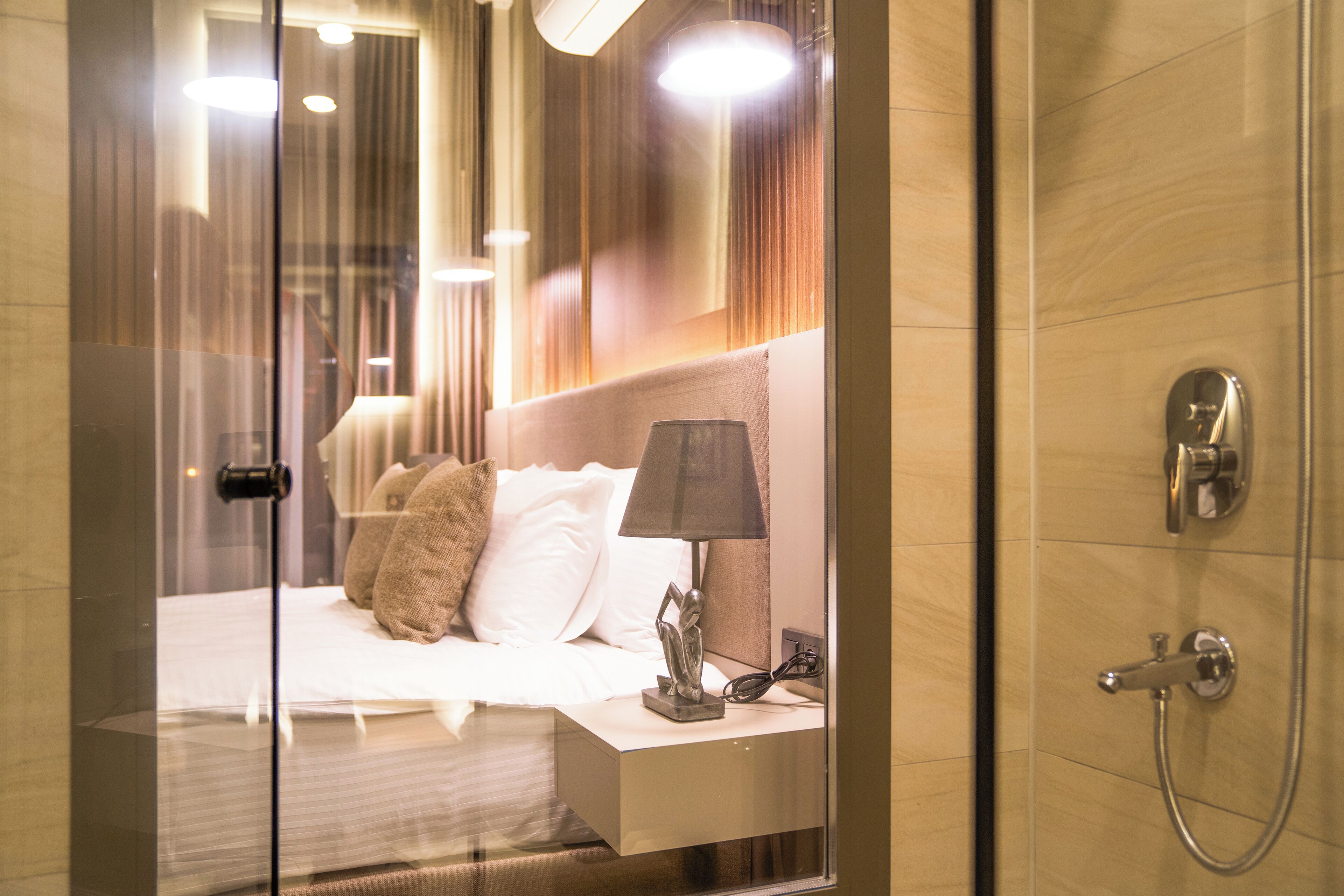 Une chambre d’hôtel moderne avec une cloison en verre, un lit soigneusement fait, une table de chevet avec une lampe et un espace douche.