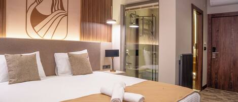 Habitación de hotel con una cama grande, cabecera de madera, mampara de ducha de vidrio y puerta de madera.