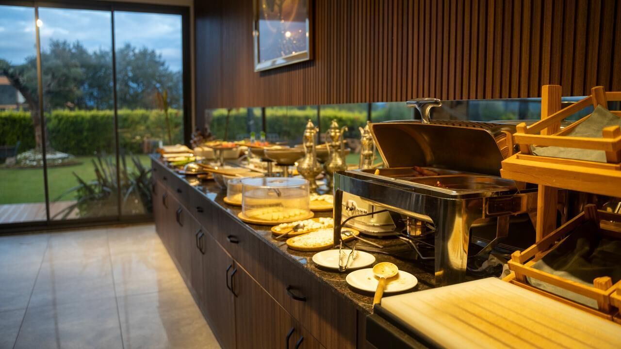 A buffet setup with various dishes and utensils, a wooden counter, and a view of greenery outside.