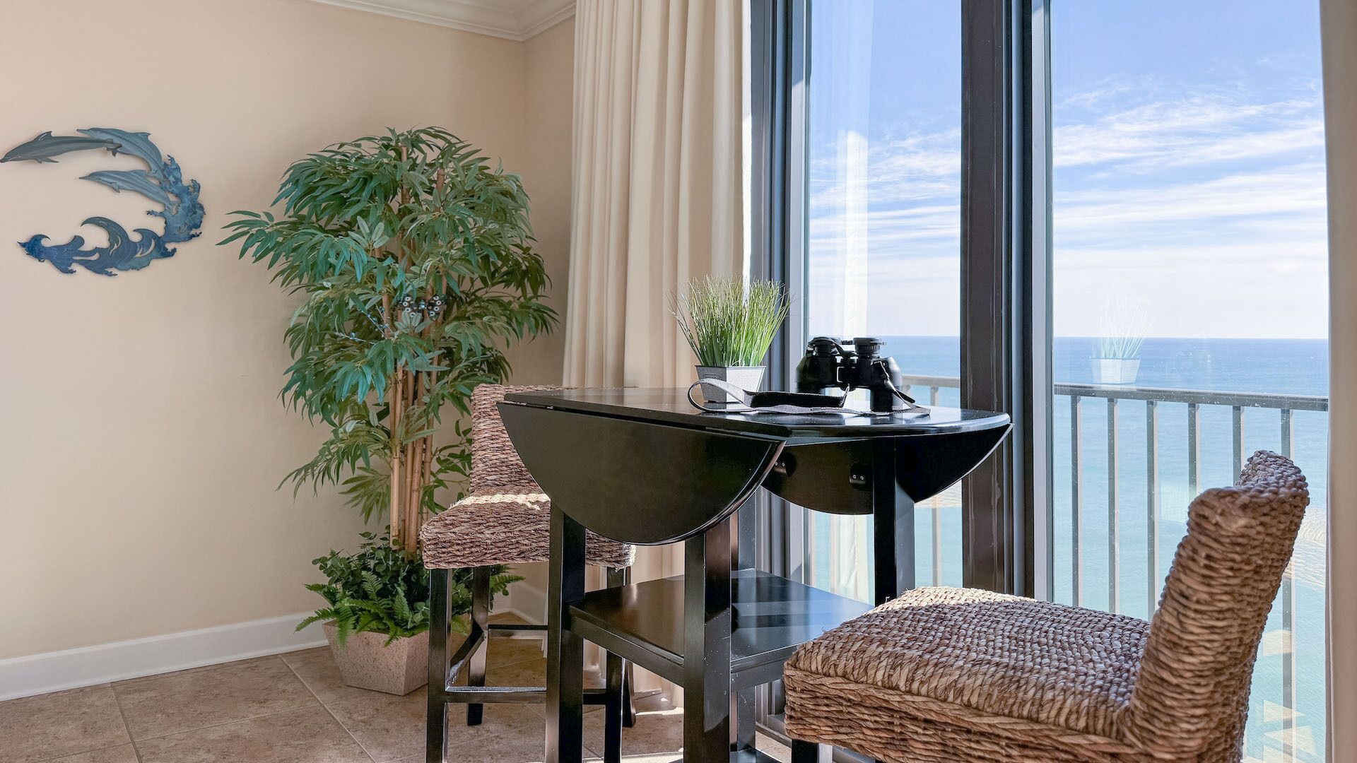 A balcony with a view of the ocean, a table with chairs, a potted plant, and a decorative wall piece.