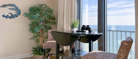 A balcony with a view of the ocean, a table with chairs, a potted plant, and a decorative wall piece.