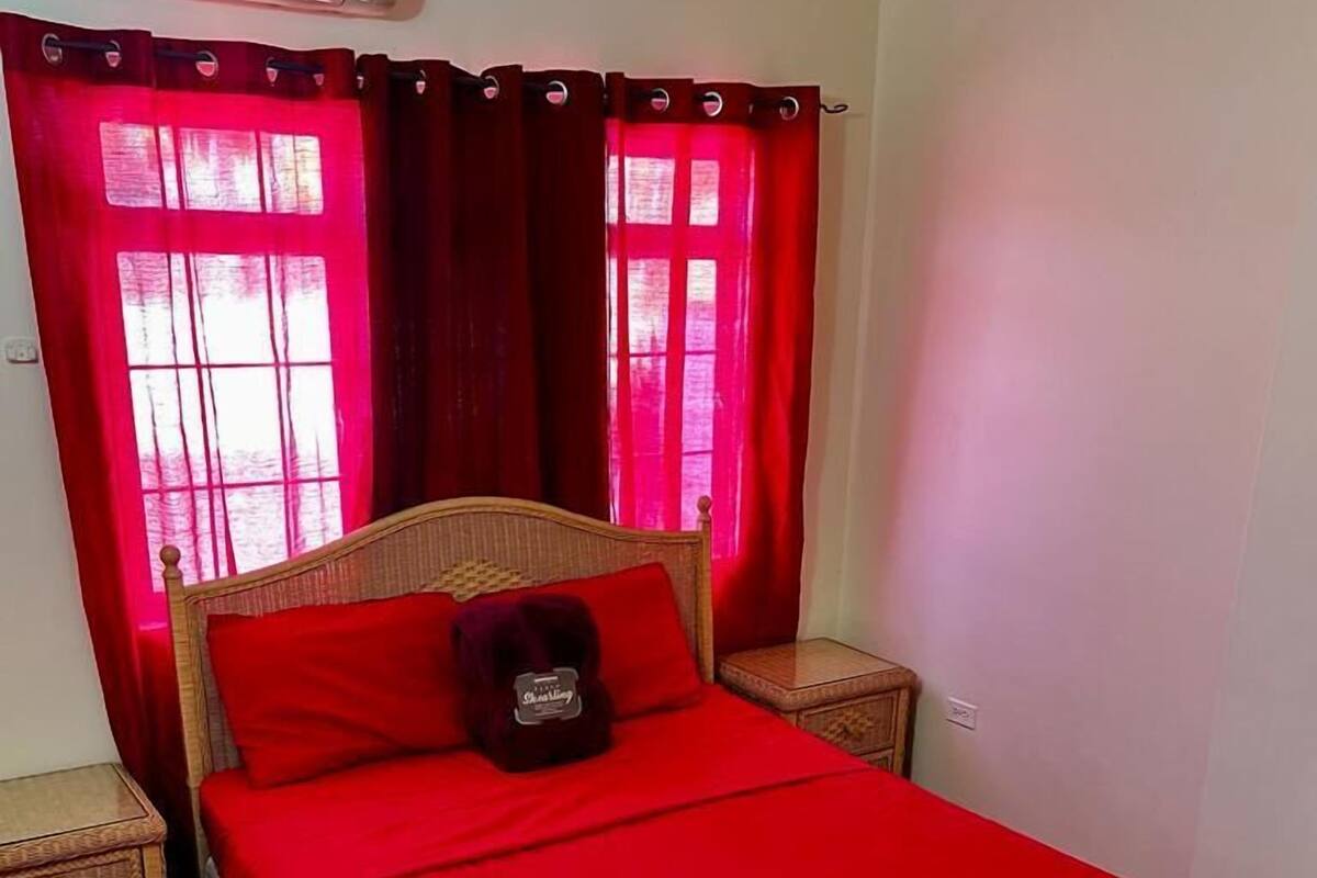 Een slaapkamer met een enkel bed, rode beddengoed, een houten hoofdbord, een nachtkastje, een rood vloerkleed en een raam met rode gordijnen.