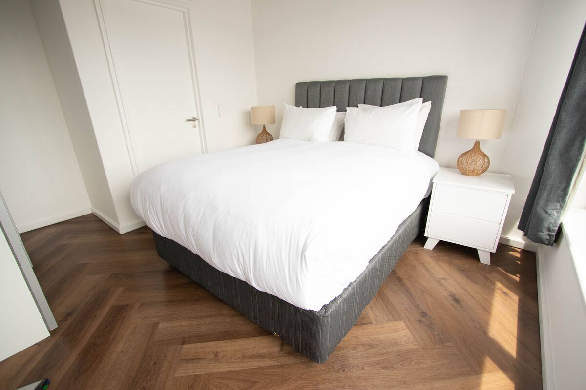 Een opgemaakt bed met een grijze hoofdbord, witte beddengoed en twee nachtkastjes met lampen.
