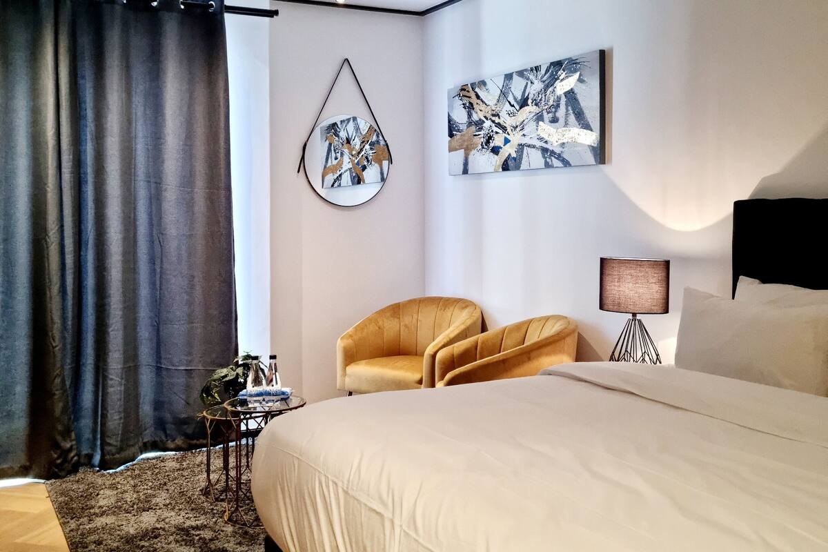 Een moderne slaapkamer met een groot bed, twee fauteuils, een hanglamp en abstracte wanddecoratie.