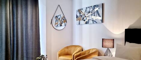 A modern bedroom with a large bed, two armchairs, a hanging light fixture, and abstract wall art.
