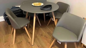 A corner room with a round table and four grey chairs. A plate of food is on the table, and a black bag is on the floor. - Captivating 1-bed Apartment in Stratford (London)