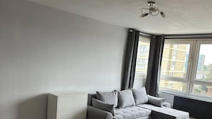 A modern living room with a grey sectional sofa, a white radiator, and a large window with curtains. - Captivating 1-bed Apartment in Stratford (London)
