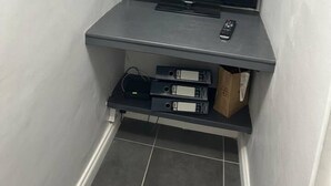 A corner with a flat-screen TV on a grey shelf, a remote control, and some electronic devices on the lower shelf. - Captivating 1-bed Apartment in Stratford (London)