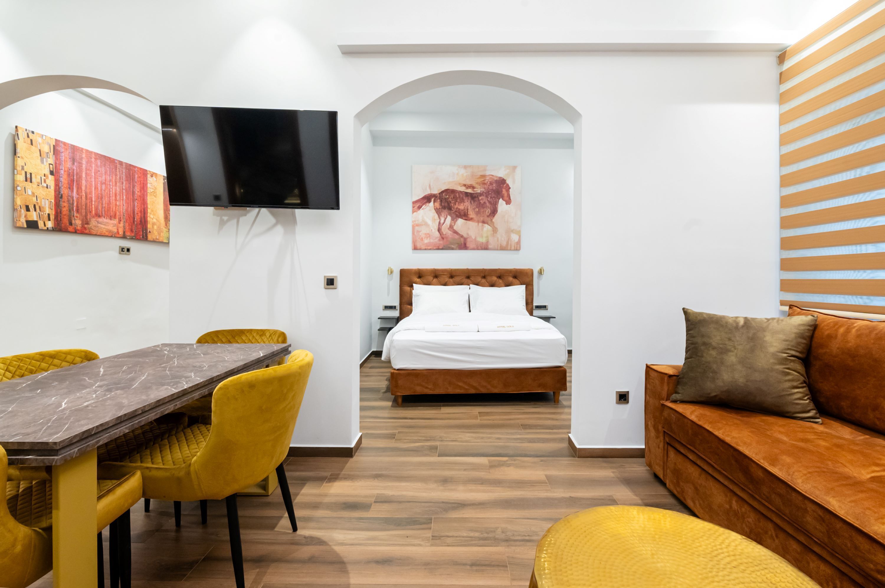 A modern hotel room with a wooden floor, a bed with white linens, a brown sofa, a dining table with yellow chairs, and artwork on the walls.