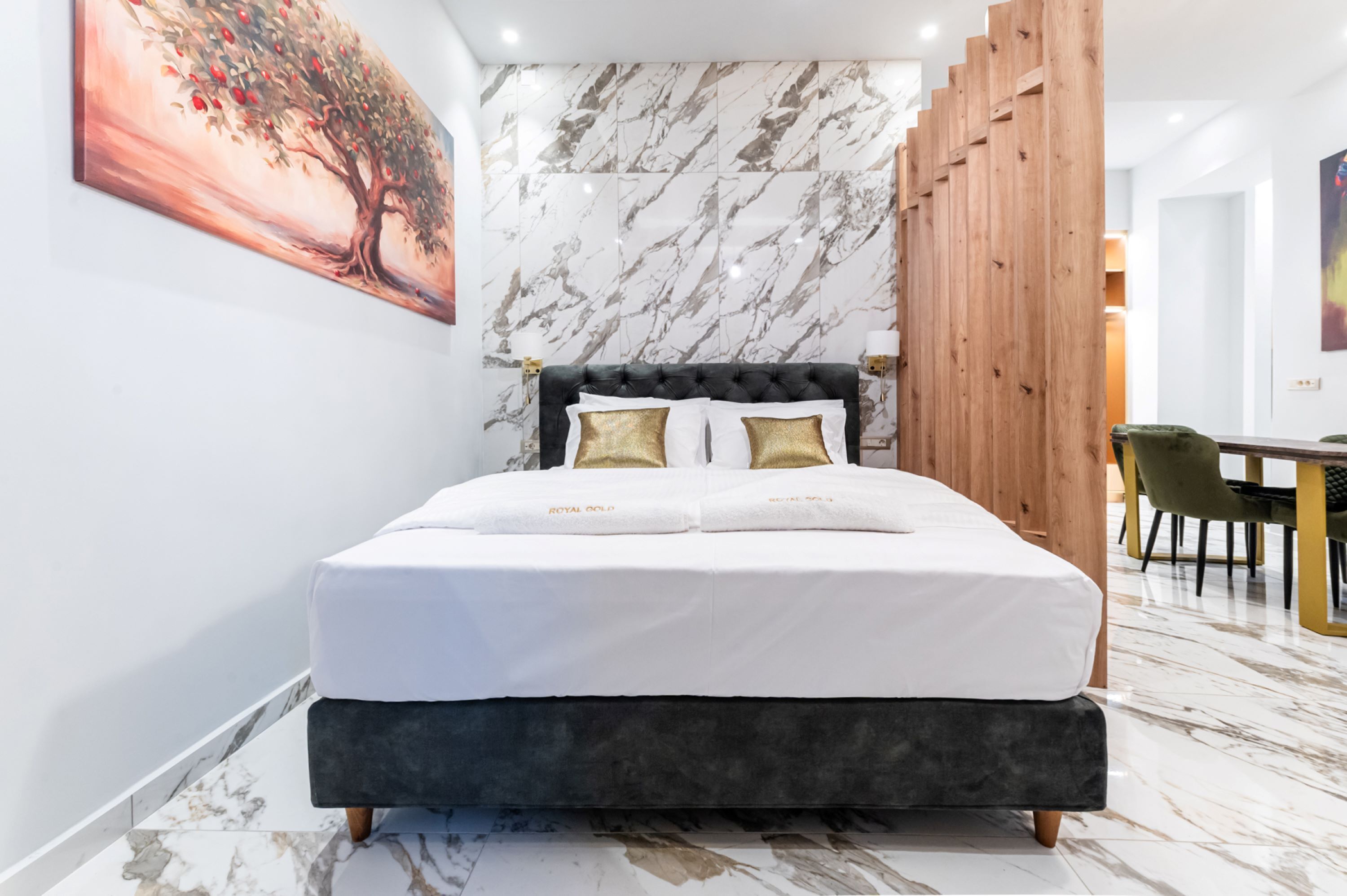 A modern bedroom with a marble-patterned wall, a tufted headboard, and a dining area in the background.