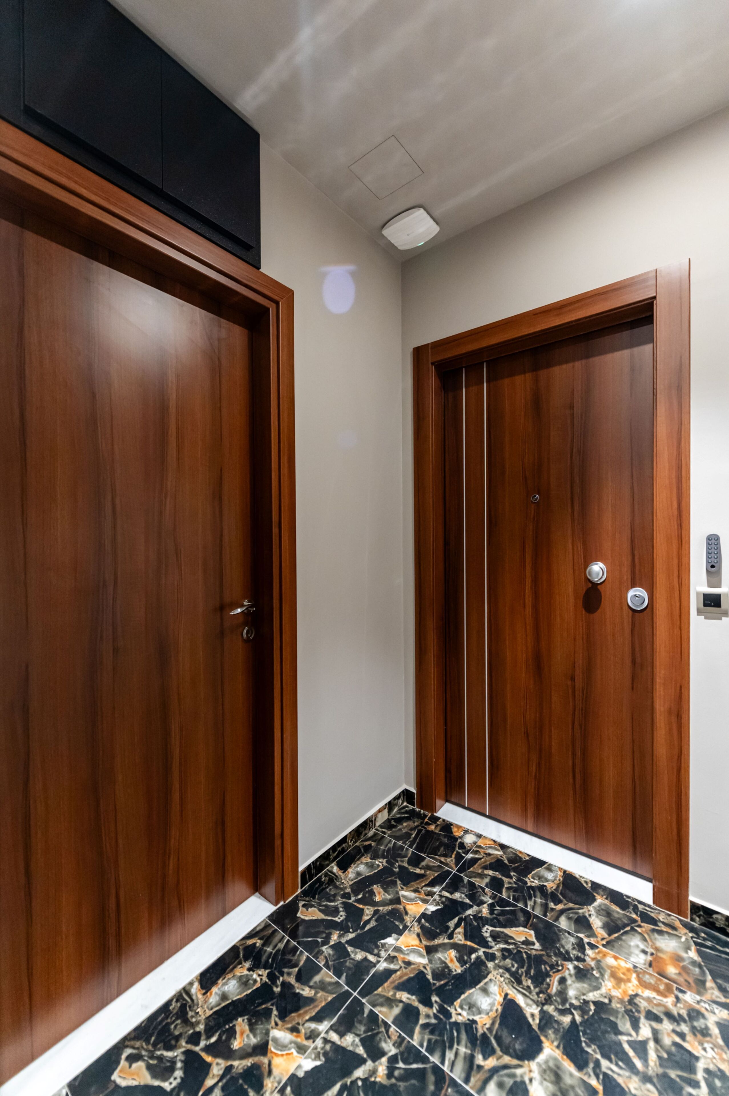 Un couloir avec deux portes en bois, un sol en marbre et un luminaire au plafond.
