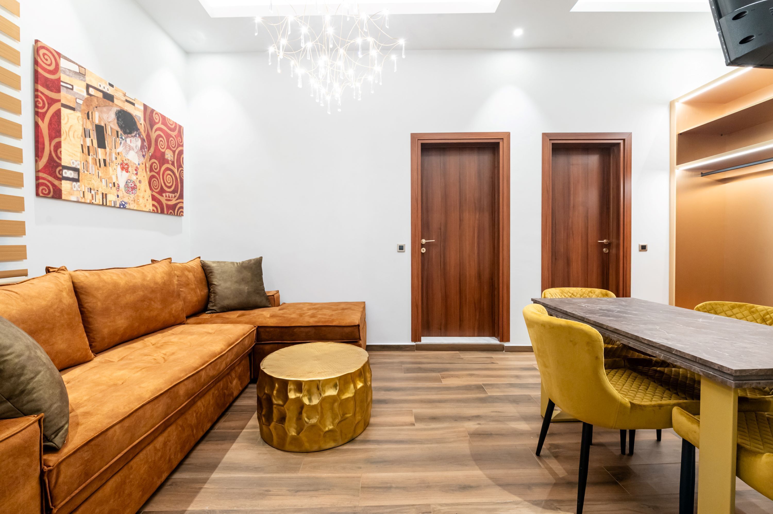 Un salon moderne avec un canapé, une table ronde dorée et des chaises jaunes. On y trouve une porte en bois et un tableau accroché au mur.