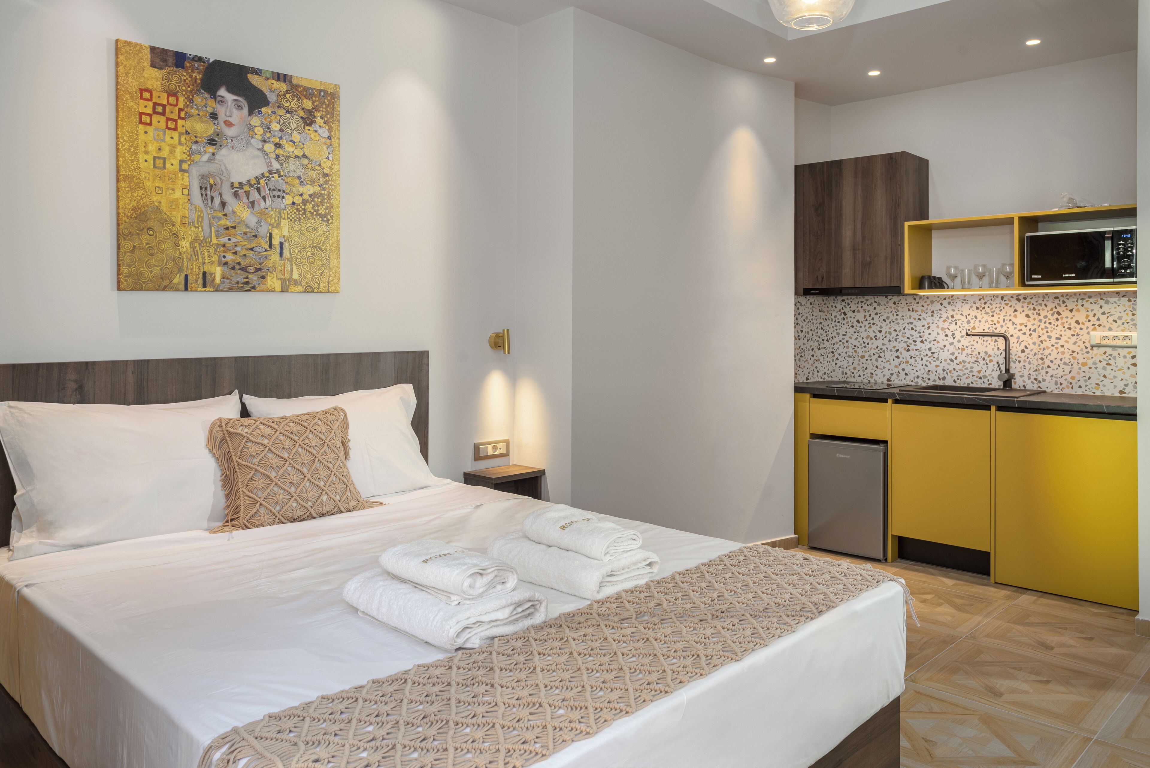 A modern hotel room with a neatly made bed, a kitchen area with yellow cabinets, and a wall-mounted artwork.