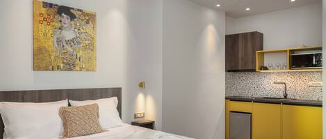 A modern hotel room with a neatly made bed, a kitchen area with yellow cabinets, and a wall-mounted artwork.