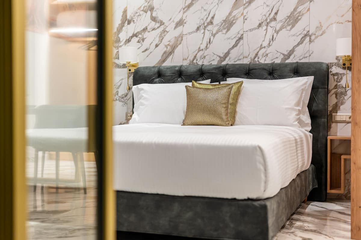 Una cama con cabecera oscura, ropa de cama blanca y un cojín de color dorado.
