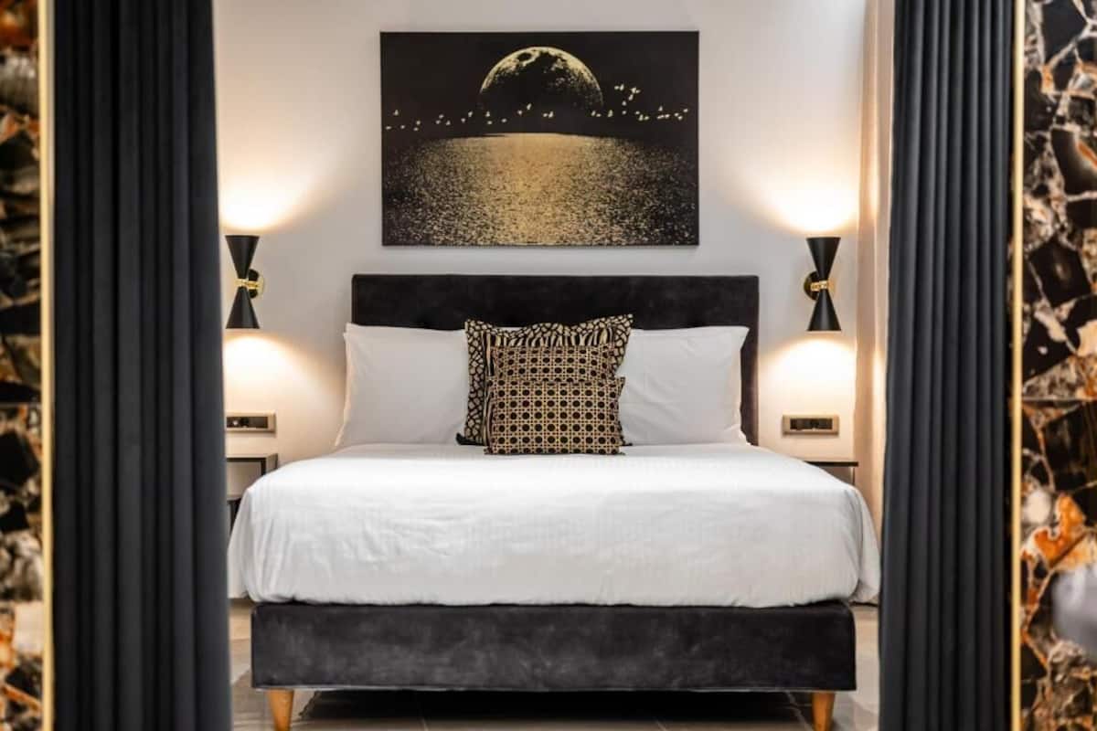 Una cama bien hecha con sábana blanca y almohada estampada, con dos lámparas fijadas a la pared a cada lado, y una obra enmarcada que muestra un paisaje iluminado por la luna encima.