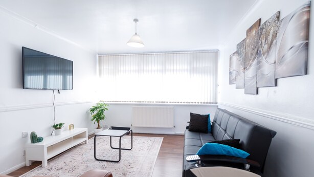 A modern living room with a black sofa, a white coffee table, a TV mounted on the wall, and a large window with blinds. - Inviting 3-bed House in London, Stay up to 6 (London)