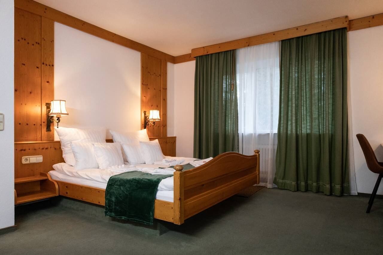 Habitación de hotel con cama de madera, dos lámparas de noche, una manta verde y una silla.