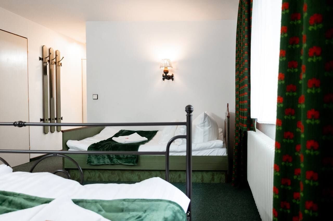 A single-bed room with a metal frame bed, a wall-mounted light fixture, a radiator, and a window with curtains.