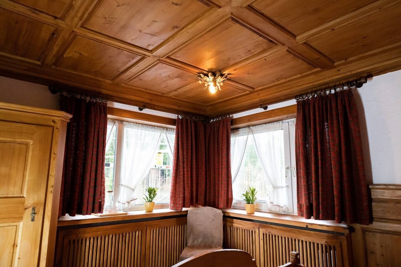 Une pièce avec un plafond et des murs en bois, deux fenêtres avec des rideaux rouges, un radiateur et une chaise.