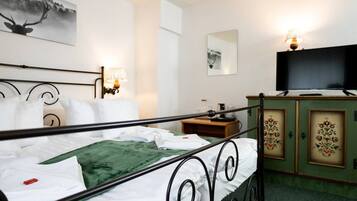 A neatly made bed with white linens and a green blanket, a television on a wooden cabinet, and wall art including a deer and a landscape.