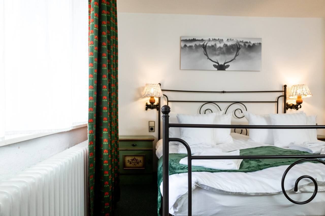 Un lit bien fait, avec du linge de lit blanc et un plaid vert, une table de chevet avec une lampe, un tableau encadré au mur et un radiateur.