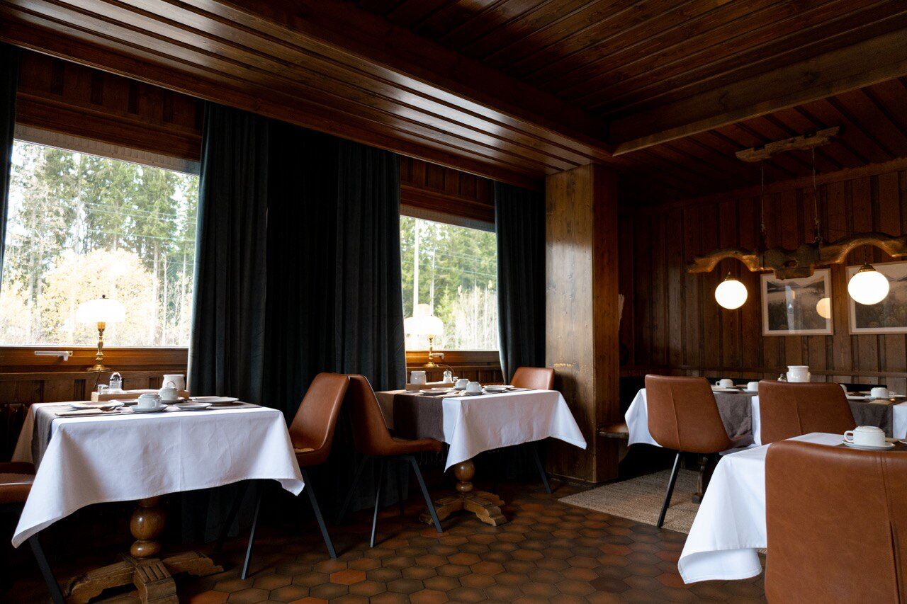 Ein Restaurant mit Holzeinrichtung, Tischen mit weißen Tischdecken und großen Fenstern mit Blick auf Bäume.