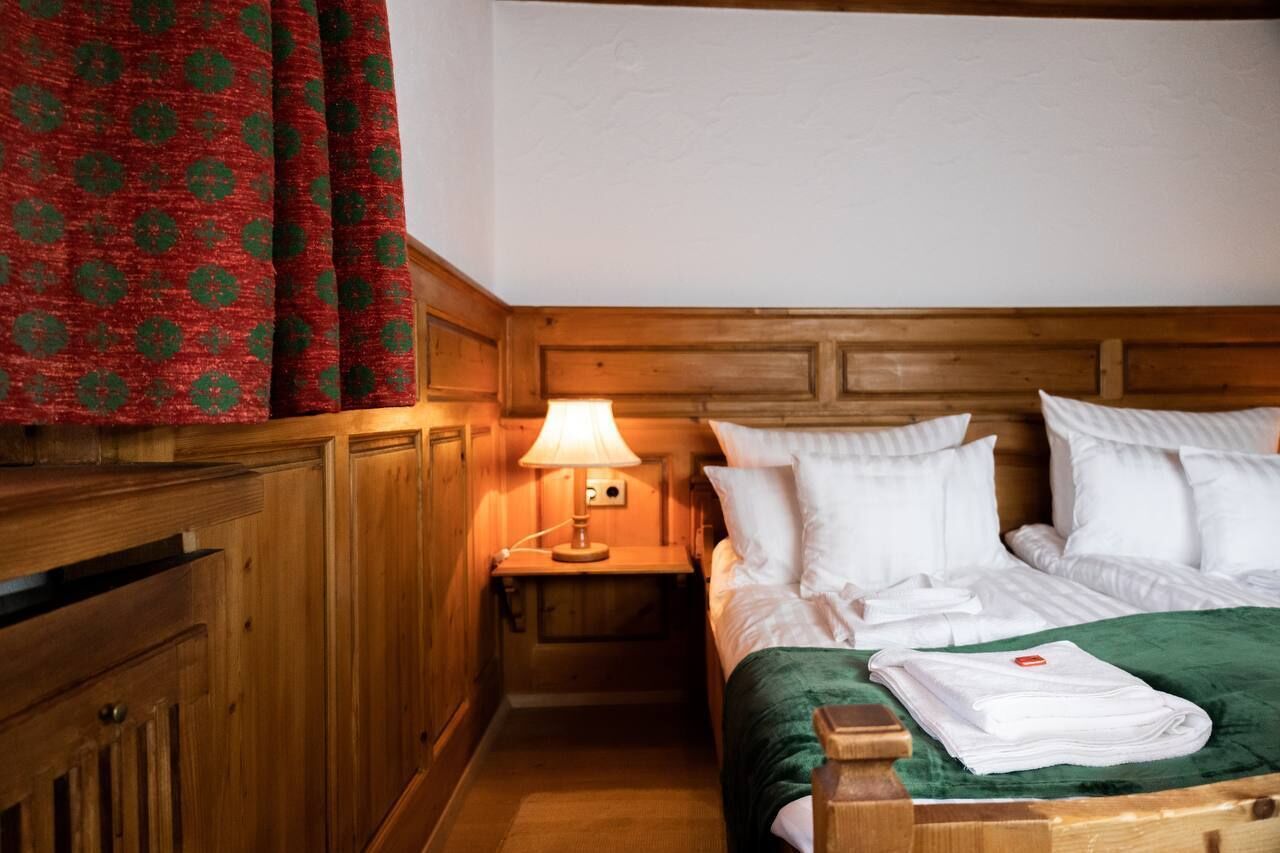 A hotel room with a wooden headboard, a neatly made bed with white linens, a green throw blanket, and a red patterned curtain.