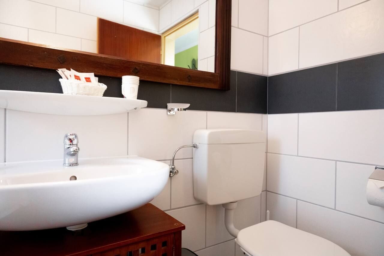 Un baño con un lavabo blanco, un espejo, un inodoro y un estante con artículos de aseo.