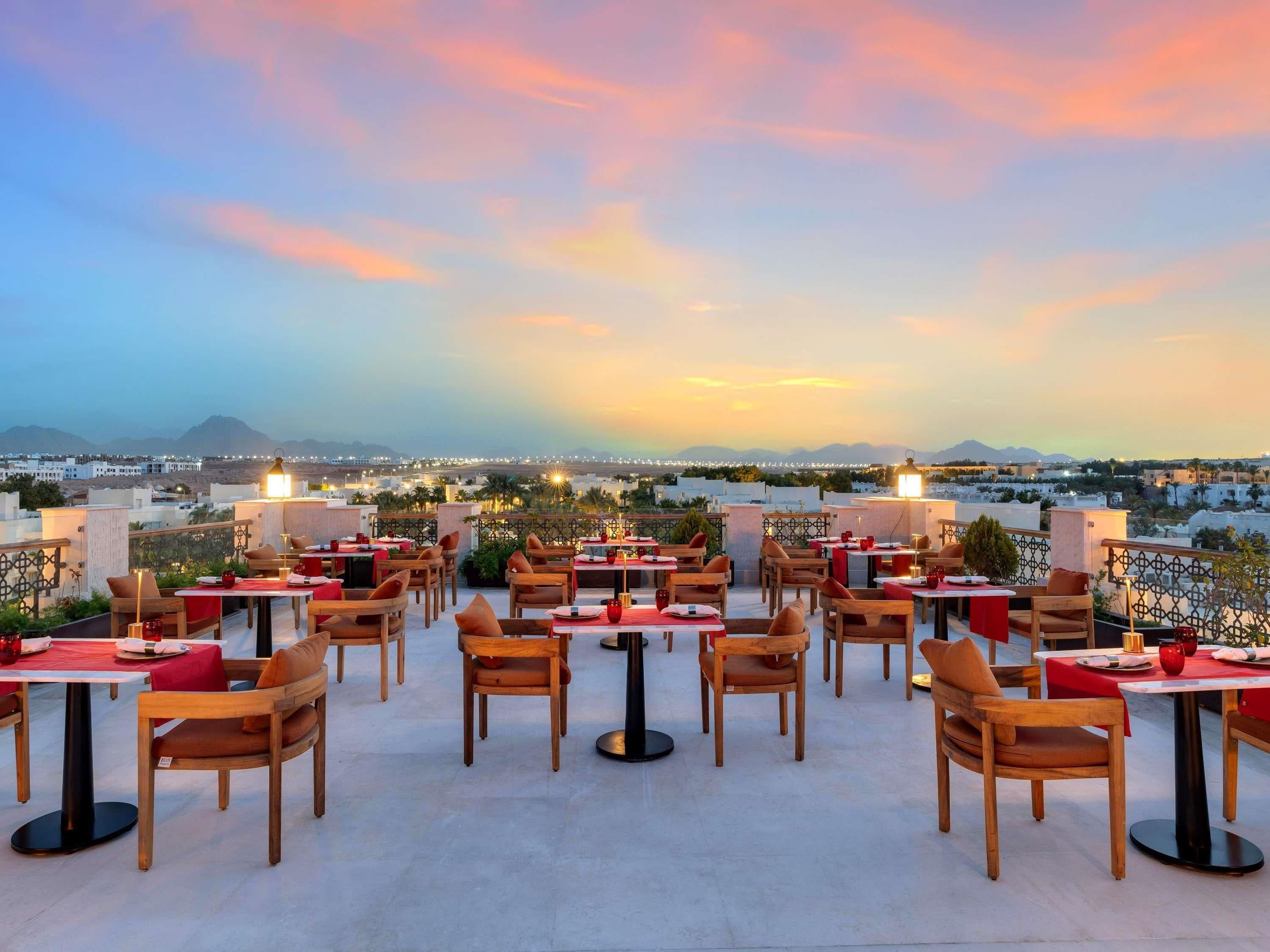 A rooftop restaurant with multiple tables and chairs, set against a backdrop of a cityscape and mountains during sunset.
