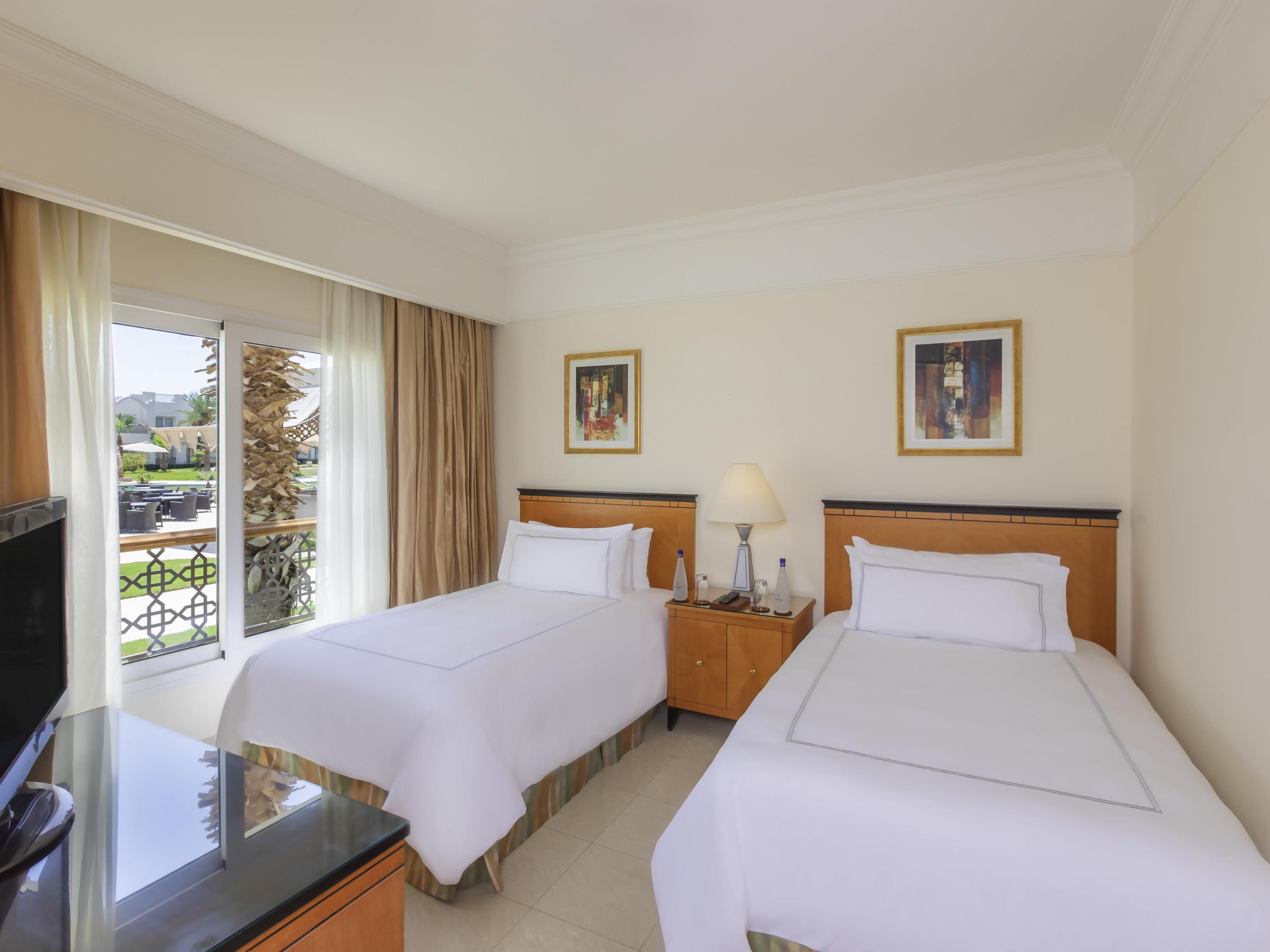 Habitación de hotel con dos camas, televisión, escritorio y balcón con vistas.