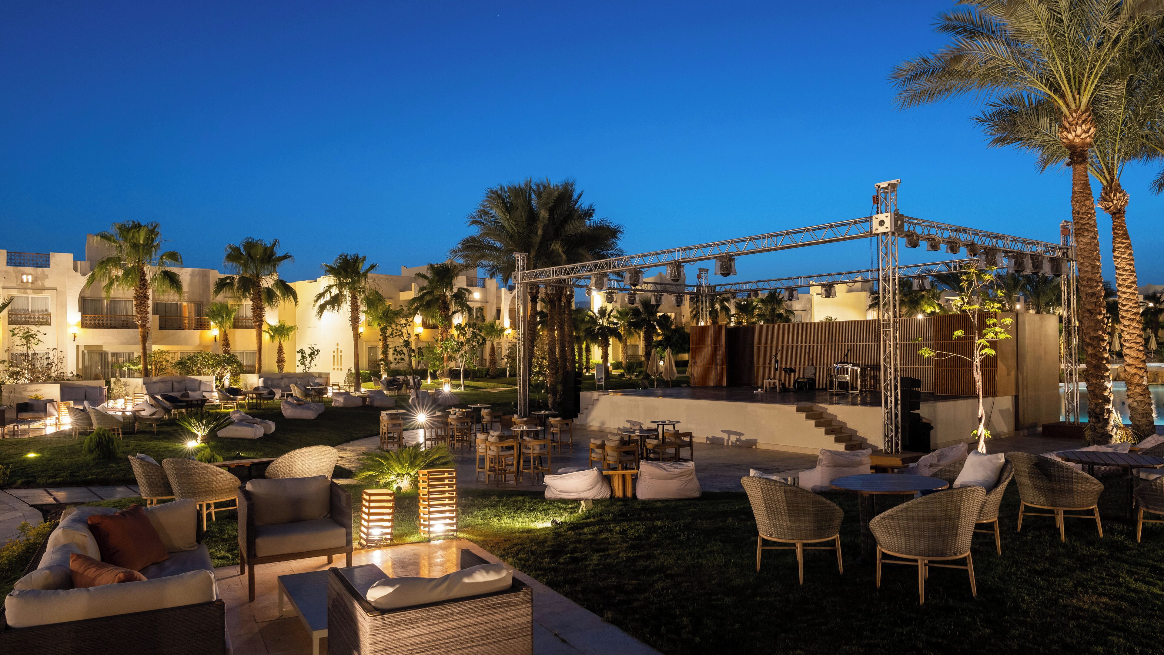 An outdoor lounge area with seating and a stage for performances, surrounded by palm trees and a pool.