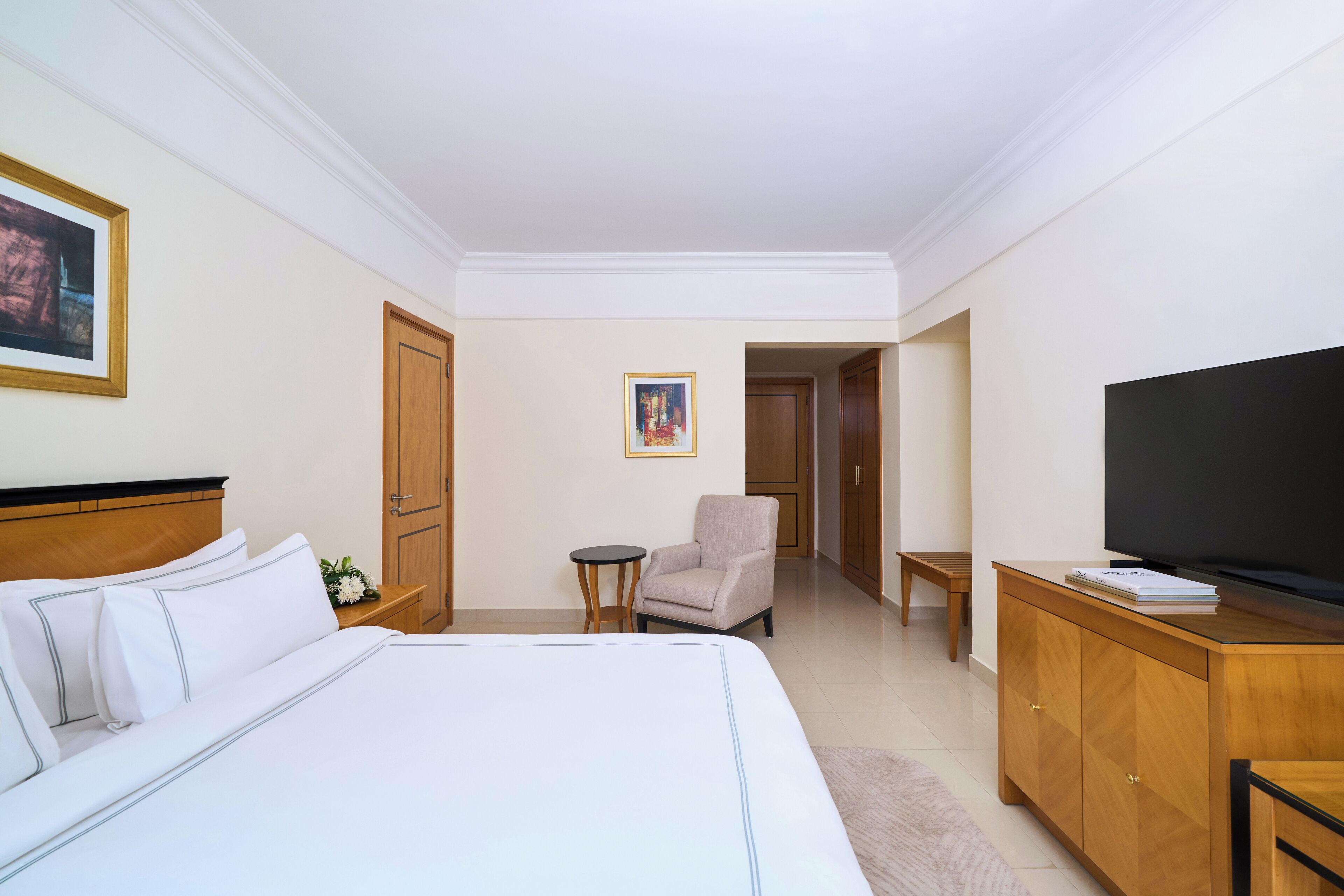 A hotel room with a large bed, a TV, a wooden cabinet, a small table, a chair, and a painting on the wall.