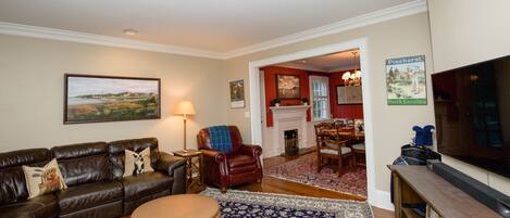 A living room with a leather couch, a round ottoman, a television, and a fireplace in the background.