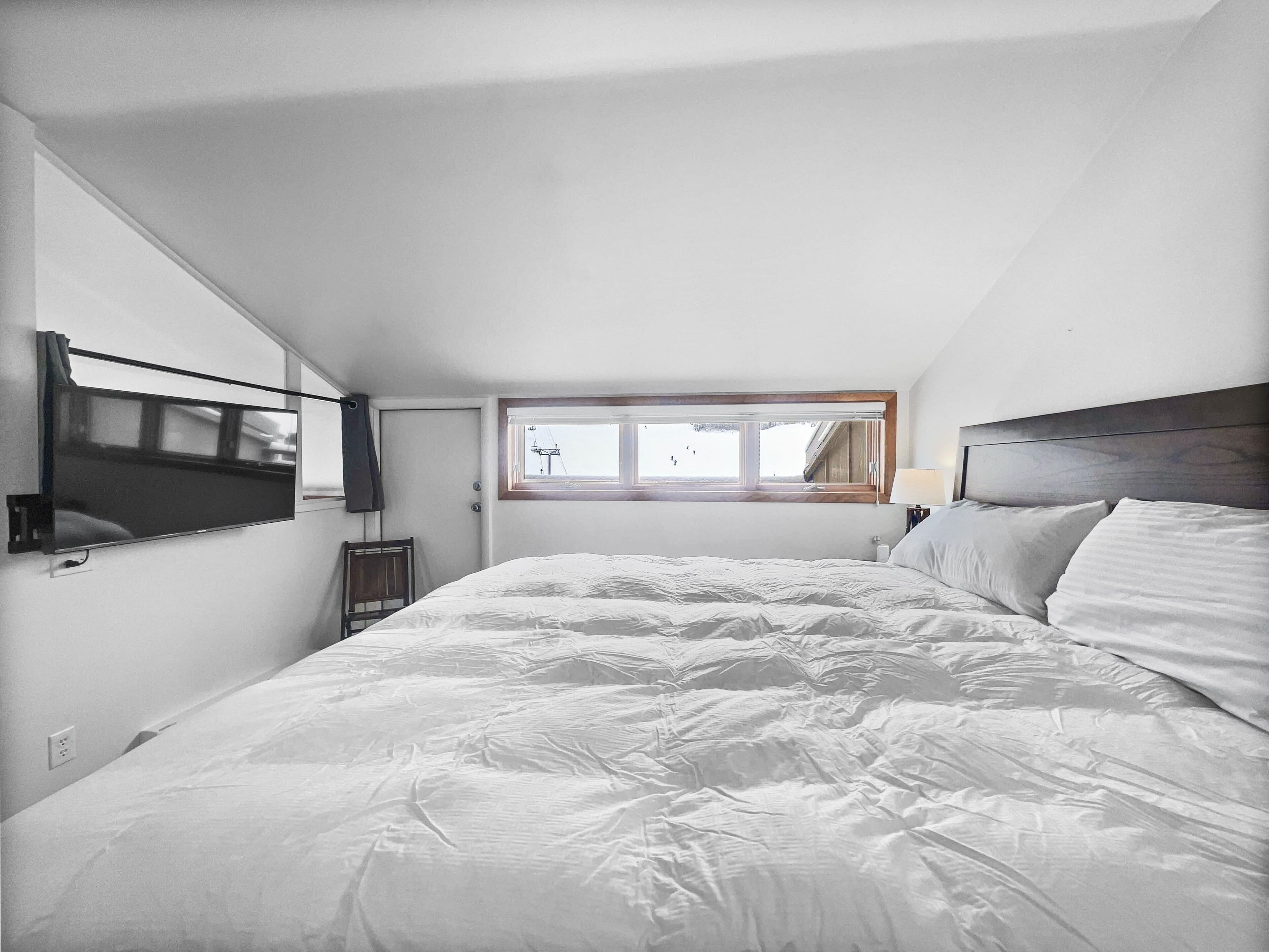 Ein Schlafzimmer mit einem großen Bett, einem flachbildfernseher an der Wand, einem Fenster mit Blick auf ein Gebäude und einem hölzernen Kopfteil.